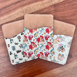 May include: Three different patterned fabric wallets with brown leather trim. The wallets feature Disney characters including Ariel from The Little Mermaid, Mickey Mouse, and Pluto.