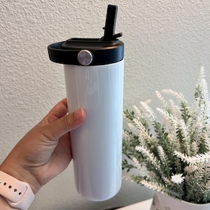 May include: A white cylindrical tumbler with a smooth, glossy finish. The container features a black plastic lid equipped with a built-in straw mechanism and a circular carrying handle accented by a small, metallic silver circular button on the side.