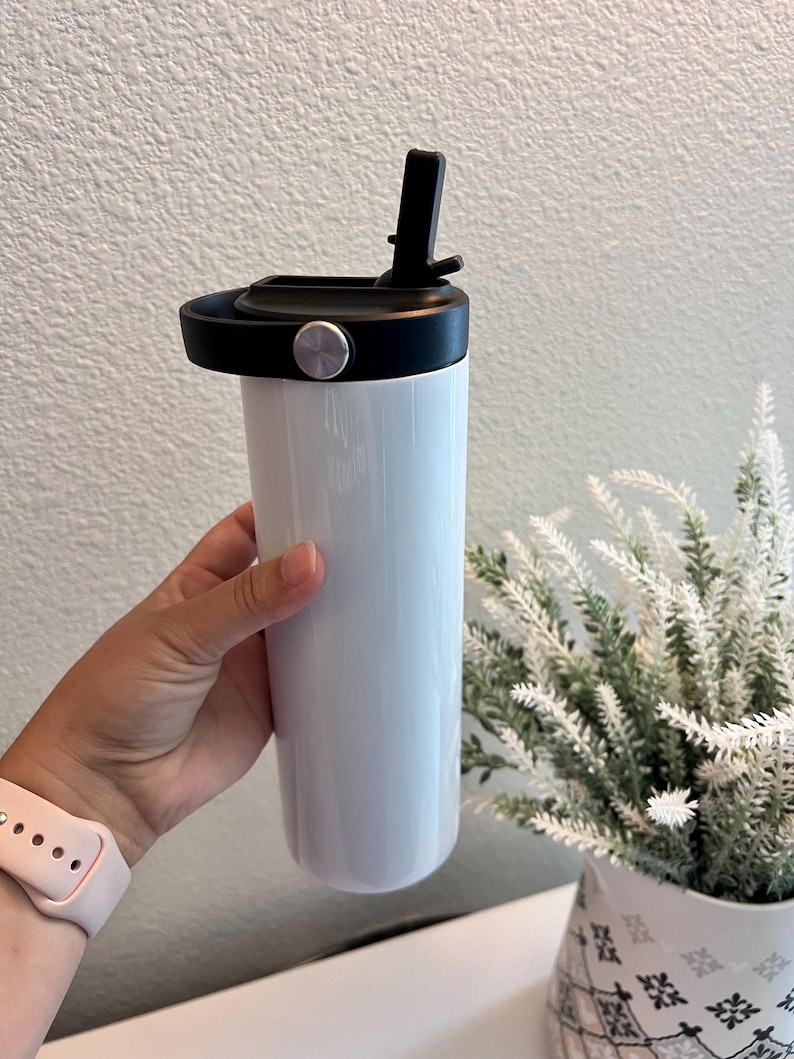 May include: A white cylindrical tumbler with a smooth, glossy finish. The container features a black plastic lid equipped with a built-in straw mechanism and a circular carrying handle accented by a small, metallic silver circular button on the side.
