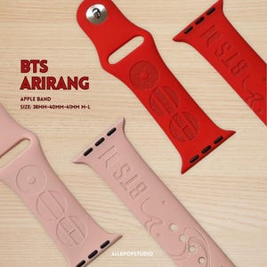Pode incluir: Pulseiras vermelhas e rosa para Apple Watch com o texto "BTS" e "ARIRANG". As pulseiras são feitas de silicone e apresentam desenhos em relevo. O tamanho é de 38 mm a 41 mm M-L. As pulseiras são exibidas em uma superfície de madeira.