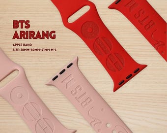 Puede incluir: Correas rojas y rosas para Apple Watch con el texto "BTS" y "ARIRANG". Las correas son de silicona y presentan diseños en relieve. El tamaño es de 38 mm a 41 mm M-L. Las correas se muestran sobre una superficie de madera.