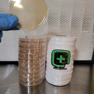 May include: A stack of clear plastic petri dishes with a single petri dish held in a blue gloved hand. The petri dish is filled with a clear, slightly yellow liquid. The stack of petri dishes is wrapped in clear plastic wrap. A white container with a green logo is next to the stack of petri dishes.