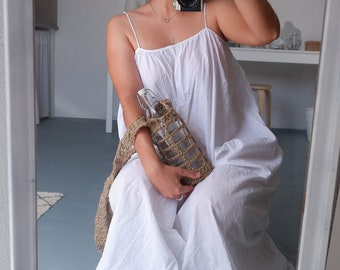 May include: A woman wearing a white sundress sits on the floor holding a glass bottle in a woven bag. She is taking a photo with a camera.