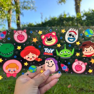 May include: A clear sticker sheet featuring colorful cartoon characters from the movie "Toy Story." The sheet includes images of Woody, Buzz Lightyear, and other characters, along with stars and planets. The text "TOY STORY" is also visible.