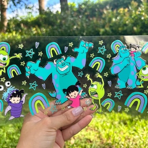 May include: A clear sticker sheet featuring cartoon characters from the movie Monsters, Inc. The sheet includes images of Sully, Mike Wazowski, Boo, and other characters, along with rainbows and stars. The characters are in shades of blue, green, purple, and pink.