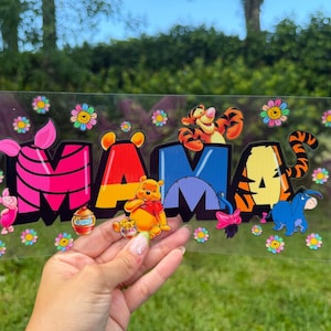 May include: A clear rectangular decal featuring the word MAMA in large block letters. Each letter incorporates Winnie the Pooh characters including Piglet, Pooh Bear, Tigger, and Eeyore, decorated with colorful flower accents throughout the design.
