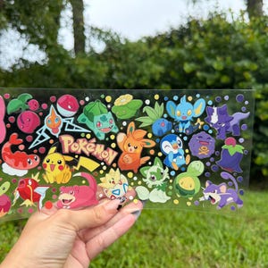 May include: A clear sticker sheet featuring a variety of colorful cartoon characters including Pikachu, Bulbasaur, Piplup, and others, with the word Pokémon printed in the center, surrounded by small circular accents in various bright colors.