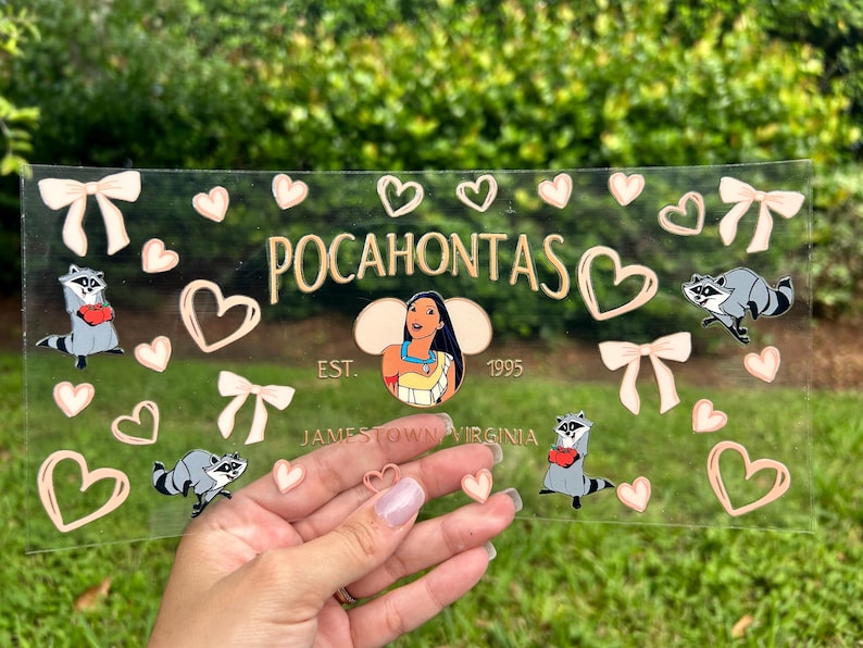 May include: Clear acrylic sign featuring the word "Pocahontas" in gold lettering, with a cartoon image of Pocahontas. The sign includes raccoon illustrations, hearts, and bows. The text "EST. 1995" and "Jamestown Virginia" are also present.