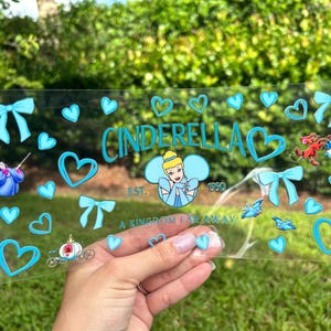 May include: A clear acrylic sign featuring the word "Cinderella" in teal, along with hearts, bows, and illustrations of Cinderella and other characters. The sign also includes the text "A Kingdom Far Away" and "Est. 1950".