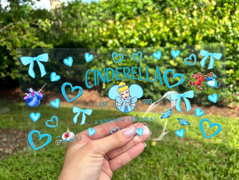 May include: A clear acrylic sign featuring the word "Cinderella" in teal, along with hearts, bows, and illustrations of Cinderella and other characters. The sign also includes the text "A Kingdom Far Away" and "Est. 1950".
