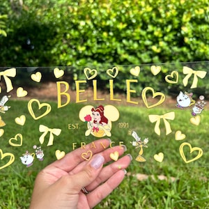 May include: A clear, rectangular decorative piece with the name "BELLE" in gold lettering, surrounded by gold hearts, bows, and character illustrations. The text "EST. 1991 FRANCE" is also present. The piece is held in a hand.