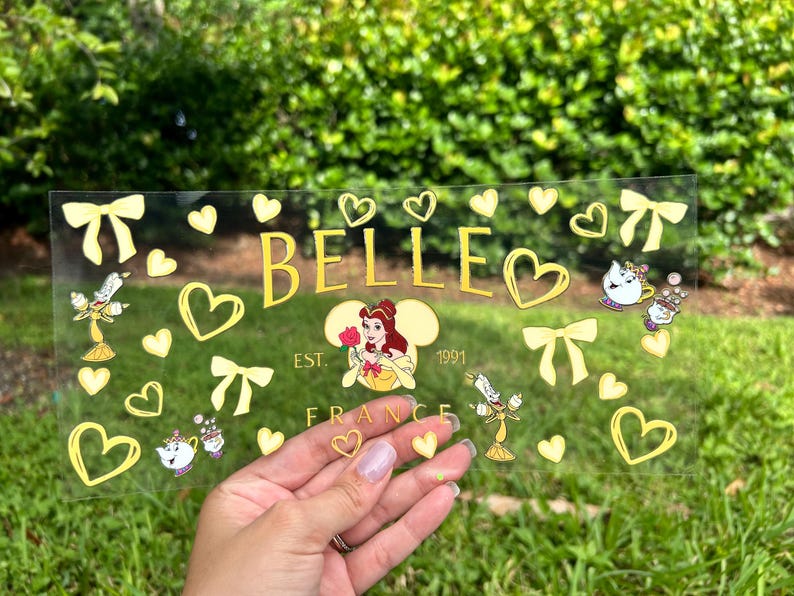 May include: A clear, rectangular decorative piece with the name "BELLE" in gold lettering, surrounded by gold hearts, bows, and character illustrations. The text "EST. 1991 FRANCE" is also present. The piece is held in a hand.