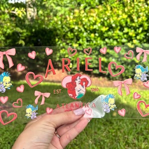 May include: A clear acrylic sign featuring the name "ARIEL" in pink, surrounded by pink hearts, bows, and cartoon characters. The sign also includes the text "EST. 1989 ATLANTICA".