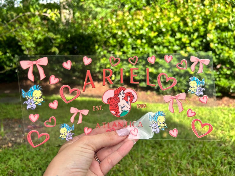 May include: A clear acrylic sign featuring the name "ARIEL" in pink, surrounded by pink hearts, bows, and cartoon characters. The sign also includes the text "EST. 1989 ATLANTICA".