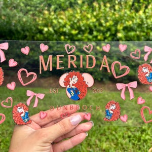 May include: A clear rectangular decorative piece with the name "MERIDA" in pink, surrounded by pink hearts, bows, and character illustrations. The text "EST. 2012 DUNBROCH" is also visible.