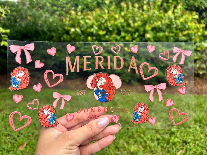 May include: A clear rectangular decorative piece with the name "MERIDA" in pink, surrounded by pink hearts, bows, and character illustrations. The text "EST. 2012 DUNBROCH" is also visible.