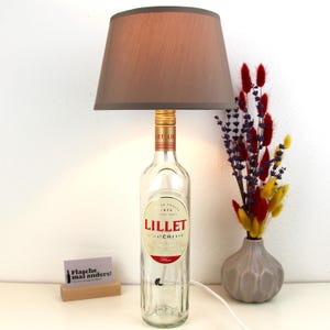 Könnte beinhalten: Eine einzigartige Tischlampe aus einer recycelten Lillet-Flasche. Die Lampe hat einen grauen Lampenschirm und einen goldfarbenen Hals. Neben der Lampe steht eine kleine Vase mit bunten Trockenblumen. Auf der Lampenbasis steht "Flasche mal anders!"