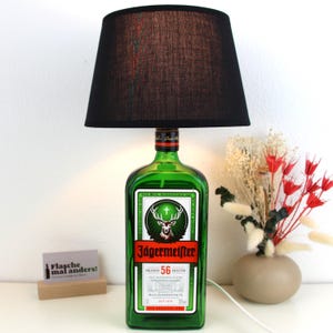 Könnte beinhalten: Eine grüne Glasflasche mit einem schwarzen Lampenschirm. Die Flasche hat ein Etikett mit der Aufschrift "Jägermeister" und zeigt einen Hirsch mit Geweih. Die Flasche steht auf einer weißen Oberfläche mit einem kleinen Holzschild, auf dem "Flasche mal anders!" steht.