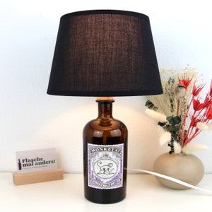 Könnte beinhalten: Ein schwarzer Lampenschirm mit einer braunen Glasflaschenbasis. Die Flasche trägt ein Etikett mit der Aufschrift "Monkey 47 Schwarzwald Dry Gin".