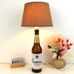 Könnte beinhalten: Ein brauner Lampenschirm mit einer Glühbirne auf einer durchsichtigen Glasbierflasche. Die Flasche ist mit "Radeberger Pilsner" beschriftet und hat ein goldenes Etikett mit einem Wappen. Die Flasche steht auf einer weißen Oberfläche mit einem kleinen Holzschild, auf dem "Flasche mal anders!" steht, und einer kleinen Vase mit Trockenblumen.