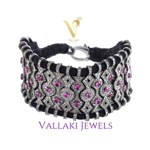May include: A silver bracelet with a black cord and a geometric design. The bracelet is adorned with small diamonds and pink gemstones. The bracelet is made by Vallaki Jewels.