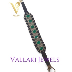 Può includere: Un braccialetto in velluto nero con un design in argento ed smeraldi. Il braccialetto presenta una chiusura con un gancio decorativo. Il testo "VALLAKI JEWELS" è visibile sul braccialetto.