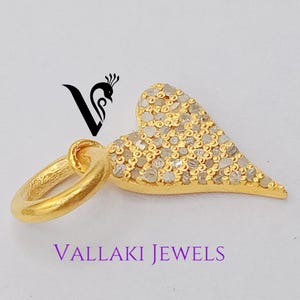Puede incluir: Un colgante de corazón de oro con un anillo para sujetarlo a una cadena. El corazón está cubierto de pequeñas piedras brillantes.