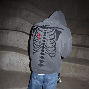 May include: A gray hooded sweatshirt with a black skeleton graphic on the back. The skeleton has a red cross on its chest.