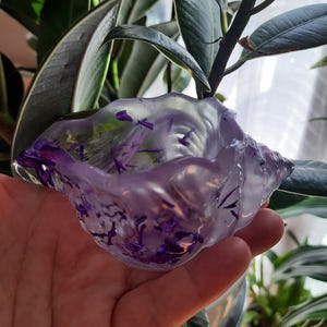 Peut inclure: Un petit plat en forme de coquille de couleur violette transparente avec des paillettes violettes incrustées dans la résine. Le plat est tenu dans la main d'une personne.