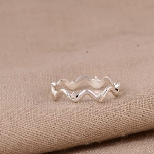 Può includere: Un anello color argento con un design ondulato. L'anello presenta un motivo continuo di picchi e valli, creando un aspetto strutturato. L'anello è esposto su un tessuto beige.