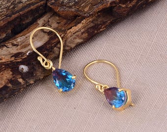 May include: A pair of gold-colored drop earrings. Each earring features a teardrop-shaped, multi-colored gemstone with hues of blue and purple. The earrings are displayed on a neutral-colored surface with a wooden background.