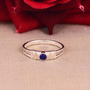 Puede incluir: Un anillo de plata con una piedra preciosa azul oscuro, redonda y central. El anillo presenta un diseño con tres formas de media luna. El anillo está sobre un fondo neutro con una flor roja en el fondo.