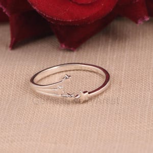 Op de afbeelding: Een zilverkleurige ring met een sterrenontwerp. De ring heeft drie kleine sterren en een gebogen band. De ring staat tegen een neutrale achtergrond, met een rode roos op de achtergrond.