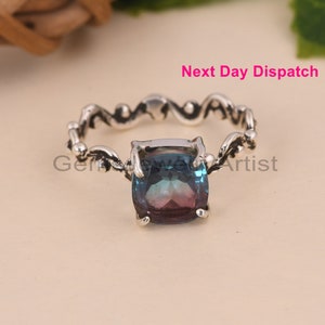 May include: A silver ring with a blue and purple gemstone set in a square shape. The ring band is ornate and features a swirling design.