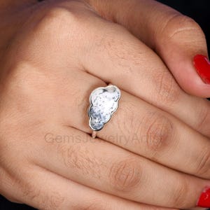 May include: A silver ring with a cloud-shaped, white gemstone with black markings. The ring is set in a silver band. The ring is shown on a hand with red nail varnish.