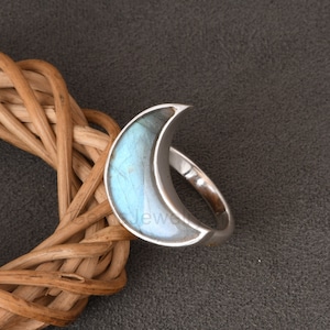 Puede incluir: Un anillo de plata con una piedra preciosa de labradorita en forma de media luna. La piedra preciosa es de color azul claro con destellos iridiscentes.