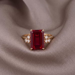 May include: A gold ring featuring a large, rectangular, emerald-cut red gemstone as the centerpiece. The band is accented on both sides by a cluster of three small, round, clear white stones, creating a balanced and symmetrical design.