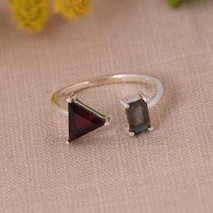 May include: A silver ring with a triangular, deep red gemstone and a rectangular, grey-green gemstone. The open design ring is displayed on a neutral surface, highlighting the contrasting colours and shapes of the gemstones.