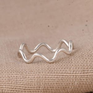 Puede incluir: Un anillo plateado con un diseño ondulado. El anillo presenta un patrón continuo y fluido, que recuerda a las olas del océano. Fabricado con un material metálico brillante, se presenta sobre una superficie texturizada de tono neutro.