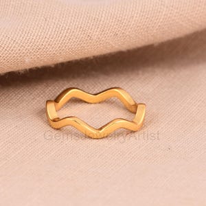 Peut inclure: Une bague dorée avec un motif ondulé continu. Le motif ondulé de la bague crée une forme fluide et organique. La bague est posée sur un fond neutre et texturé. La bague est un bijou.