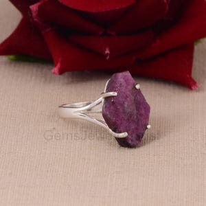 Puede incluir: Un anillo de plata con una gema grande, de forma irregular, de color magenta intenso. La gema está montada sobre una banda de vástago dividido. El anillo está sobre un fondo neutro con una rosa roja en el fondo.