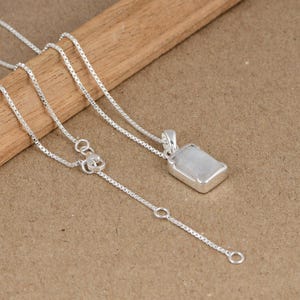 May include: A silver necklace with a rectangular, light-colored pendant. The pendant is set in a silver frame and hangs from a delicate chain. The chain has an adjustable length. The necklace is displayed on a wooden surface.