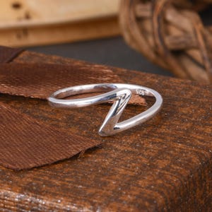 以下が含まれることがあります： モダンでミニマリストなデザインのスターリングシルバーリング。リングは滑らかで磨かれた仕上げで、特徴的なジグザグの形をしています。リングは茶色の背景に配置されています。