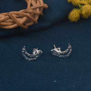 Pode incluir: Um par de brincos de prata em forma de lua crescente. Cada brinco apresenta uma textura martelada e um design curvo. Os brincos são exibidos em um tecido azul escuro.