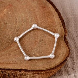 May include: A silver ring featuring a pentagonal shape formed by five thin, smooth metal bars connected at each corner by a small, polished silver sphere. The design creates a geometric, open-frame structure with a bright, reflective metallic finish.