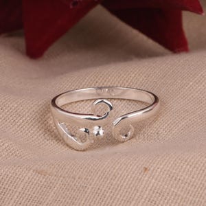Könnte beinhalten: Ein silberfarbener Ring mit offenem Design. Der Ring hat ein wirbelndes, durchbrochenes Muster mit einem kleinen, runden Akzent. Der Ring ist vor einem neutralen Hintergrund platziert.