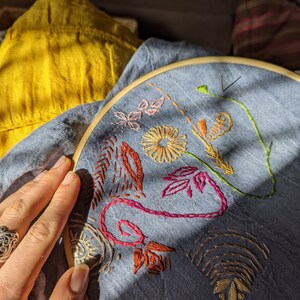 Può includere: Primo piano di un cerchio da ricamo in tessuto blu con disegni di filo colorato di fiori, foglie e spirali. Il cerchio è tenuto da una mano che indossa un anello.