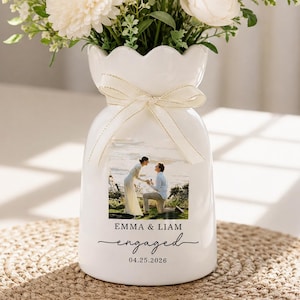 May include: A white ceramic vase with a scalloped rim and a cream-colored ribbon tied around the neck. The front features a printed photograph of a couple, with the text EMMA & LIAM, engaged, and the date 04.25.2026 printed in black ink.