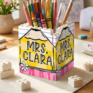 May include: A square pencil holder constructed from white building blocks, featuring a yellow pencil graphic with the text "MRS. CLARA" in black, accented by pink stripes and white flowers, filled with various colored pencils and a clear pen.