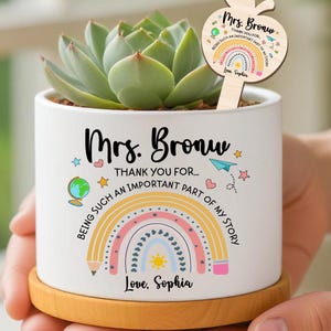 May include: A white ceramic planter with a wooden base, holding a succulent plant. The planter is decorated with a rainbow design and the text "Mrs. Bronw, Thank you for being such an important part of my story." An apple-shaped wooden stake is in the plant.